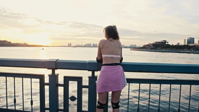 Female skater rides to railing watching passing boat on river. Beautiful young woman takes break from urban rollerblading session to admire scenic sunset.