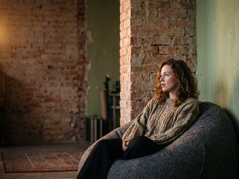 Thoughtful young woman relaxing in a beanbag chair inside a cozy rustic loft apartment with exposed brick walls