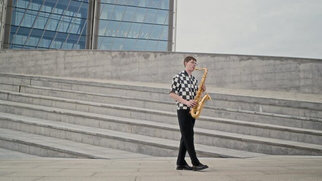 Caucasian young man playing saxophone while walking across modern urban plaza. Clip suitable for music education, cultural campaigns, street art projects, and creative industry advertising