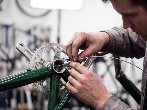 Professional bicycle mechanic threading and installing gear cable into a green bicycle frame in repair workshop