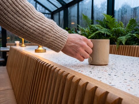 Person wearing a warm knitted sweater holding a ceramic coffee mug on a terrazzo table in a cozy modern cafe greenhouse