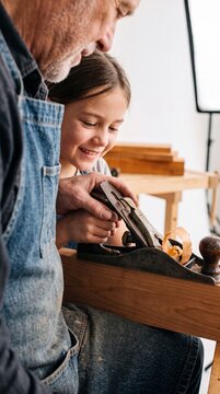 Senior carpenter teaching smiling granddaughter how to plane wood with hand plane tool in workshop