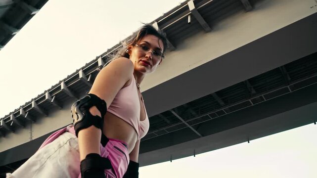Beautiful woman on roller skates stops looking directly into camera. Massive concrete bridge creates striking geometric urban lines behind confident sporty female skater outdoors.
