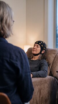 Mature woman sitting relaxed in an armchair wearing an EEG neurofeedback cap during a therapy session with a doctor