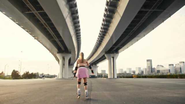 Female skater rides away into soft background focus under bridge. Active young woman wearing protective gear enjoys urban skating session during bright evening sunlight at extreme park.