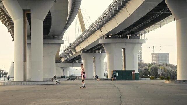 Distant view captures skater crashing during street training. Attractive sporty lady makes mistake and loses balance practicing active rollerblading lifestyle under giant city bridge structures.