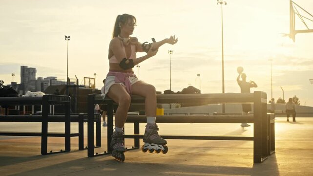 Low angle shot shows skater girl attaching black elbow protection. Active young woman prepares for outdoor rollerblading while people play basketball in background during golden sunset