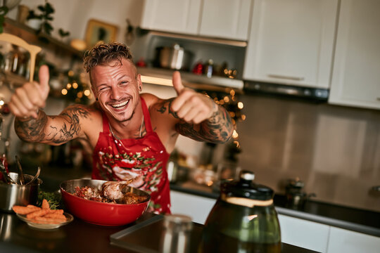 Homme souriant en tablier rouge dans une cuisine d&eacute;cor&eacute;e pour No&euml;l, pr&eacute;paration d&rsquo;un repas festif, plat de viande, cuisine familiale, convivialit&eacute;, f&ecirc;te de fin d&rsquo;ann&eacute;e et ambiance chaleureuse