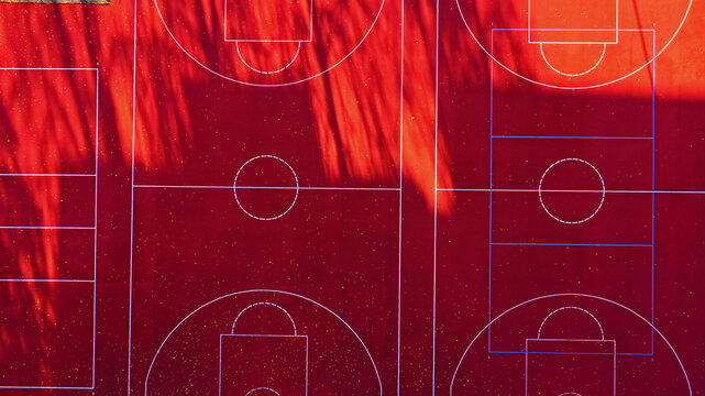 Aerial view of a vibrant red basketball court with white court markings and long tree shadows cast across the surface in Latvija.
