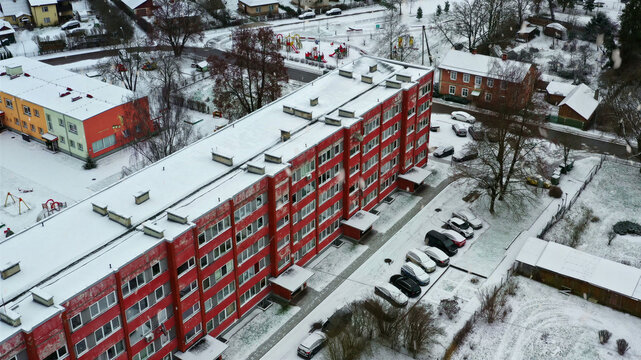 Aerial view of a red apartment building surrounded by snow-covered grounds, a parking lot, and a playground in Latvija.
