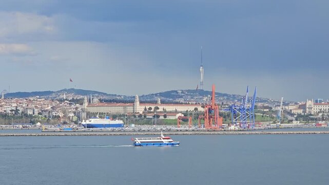 Blue ferry sails past a port with cranes and hills. Clear sky frames coastal cityscape with bridges buildings. Water reflects sky and structures in calm, steady motion in Istanbul, Turkey