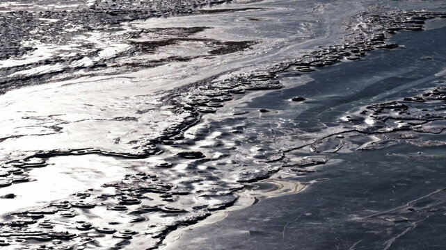 Aerial view of intricate ice patterns forming on the surface of a frozen river with reflective light and textured ice floes in Latvija.