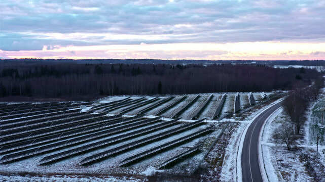 Aerial view of a solar farm with rows of photovoltaic panels covered in light snow next to a road with a forest in the background in Latvia.