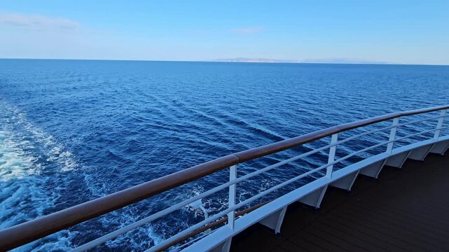 Cruise ship moves through open blue ocean. White railing lines deck edge. Wooden handrail curves beside water. Sunlight glints off calm sea surface. Blue sky stretches behind vessel