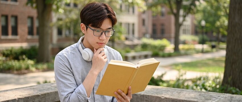 Thoughtful Asian Male Student Reading a Book Outdoors on a University Campus During Sunny Day