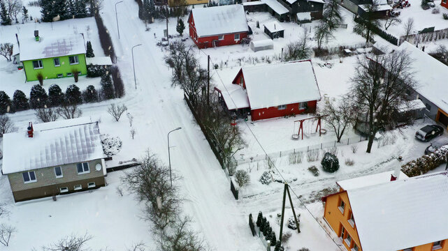 Aerial view of a quiet residential neighborhood with colorful houses surrounded by fresh snow, bare trees, and a snow-covered street in Latvija.