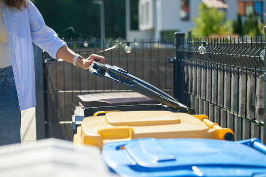 Woman opening garbage bin beside yellow and blue waste containers outside house