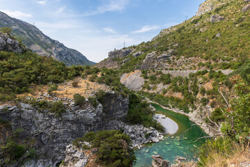 Fototapeta premium A dramatic mountain canyon scene with a turquoise Moraca river winding through rocky cliffs under blue sky. Rugged terrain and green vegetation evoke natural beauty of Montenegro