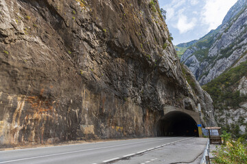 Fototapeta premium A two-lane mountain road disappears into a dark tunnel carved through a towering rocky cliff, framed by steep forested peaks and blue sky, conveying adventure, isolation and rugged engineering