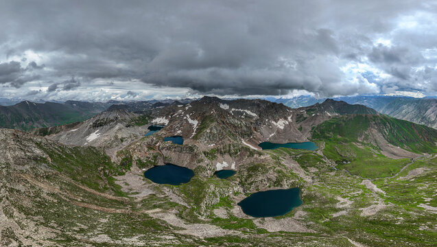 Aerial view of Maheen Lake surrounded by rocky mountain peaks, green alpine slopes, and patches of snow under a dramatic cloudy sky in Batakundi, Khyber Pakhtunkhwa, Pakistan.