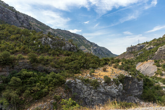 A scenic rocky canyon and valley with limestone cliffs, scrubby green hills and dry grasses beneath a clear blue sky. Rugged natural landscape suggesting hiking, geology, remote outdoors. Montenegro