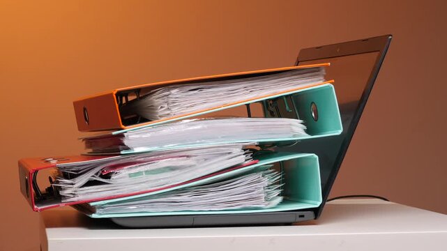 Human hands stack a towering pile of colorful binders and paperwork onto an open laptop, symbolizing office overload, digital vs. Physical tasks, stress and looming burnout