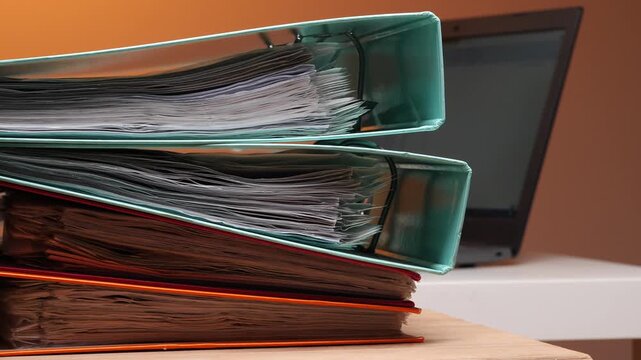Stacked ring binders overflowing with papers and reports, illustrating office paperwork overload and the need for organized filing systems on a cluttered desk with laptop background