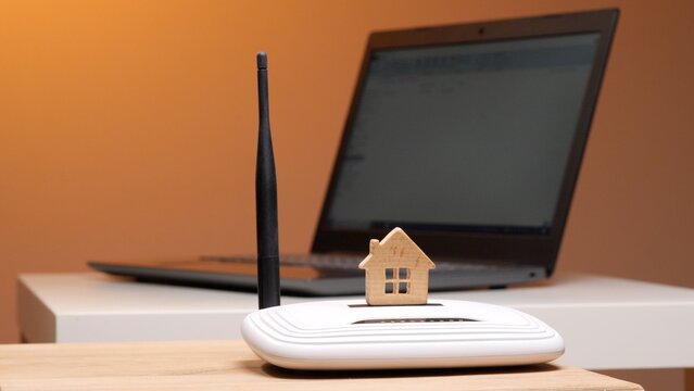 Wooden house model perched on a white wireless router beside a laptop, symbolizing smart home internet connectivity, broadband access and modern digital living on a desk