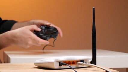 Hands holding a wired game controller with a wireless internet router connecting to the network, symbolizing online gaming, fast connectivity, and modern entertainment © Aliaksandr