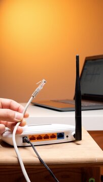 Hand holding and connecting an ethernet rj45 cable to a white wireless network router, establishing an internet connection with a laptop in the background