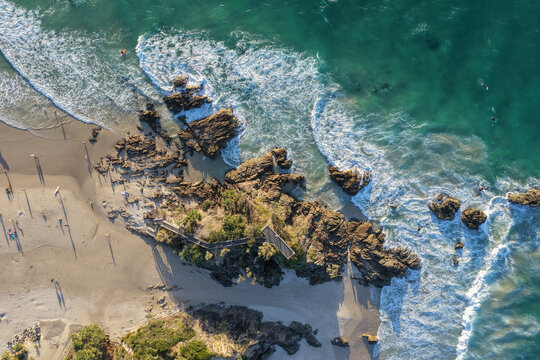 Aerial view of the rocky coastline, sandy beach, turquoise waves, and wooden lookout path at Byron Bay, New South Wales, Australia.