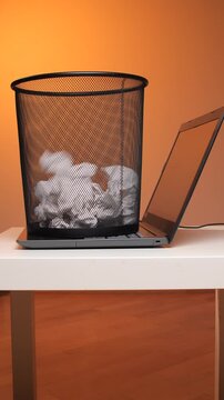 Vertical video a black mesh trash bin, overflowing with crumpled white paper, resting on an open gray laptop keyboard on a white desk, representing digital decluttering and data waste