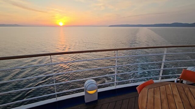 Sunset glows over calm sea from cruise ship deck. Wave trails mark ship path across tranquil waters. Golden light reflects off gentle ocean surface. Metal rail and wooden table frame the view