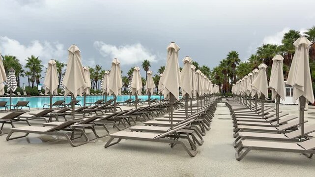 Sun loungers and closed umbrellas by resort pool on cloudy day