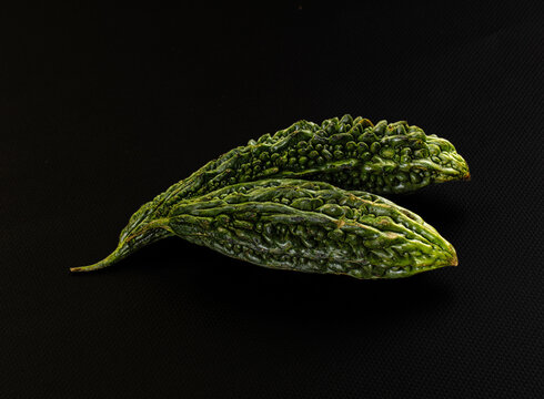 Fresh green bitter melon with warty textured skin lying on dark black fabric background, close up of raw tropical vegetable for cooking and health food