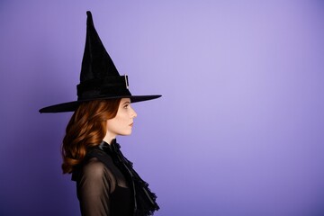 Young woman in a classic witch hat and black costume standing in profile against a purple background for Halloween portrait and fashion concept © deagreez