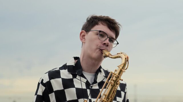 Young adult Caucasian man playing saxophone outdoors in tilt up shot. Perfect for music education, youth culture projects, festival advertising, and entertainment marketing