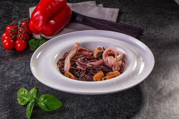 Dark slate table with squid ink spaghetti in white plate topped with octopus tentacles, mussels and cuttlefish, cherry tomatoes and pepper nearby, gourmet seafood pasta dish © Andrei Starostin