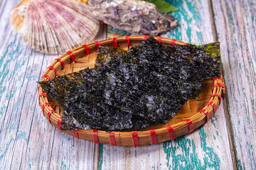 Dried nori seaweed sheets on round woven bamboo tray with red bindings and scallop shell on distressed teal wood surface © Andrei Starostin