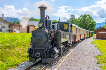 A steam locomotive is displayed at the Taurach Railway narrow-gauge museum in Austria. The train is preparing for a journey along scenic tracks. Visitors enjoy the experience. © pyty