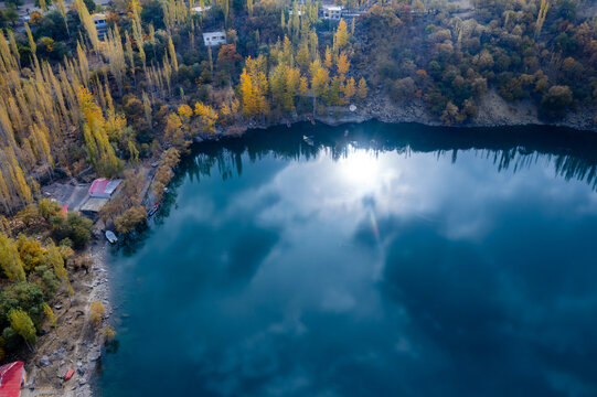Aerial view of Upper Kachura Lake with its deep blue water reflecting the sun, surrounded by vibrant autumn-colored poplar trees and rocky shores in Skardu, Gilgit Baltistan, Pakistan.