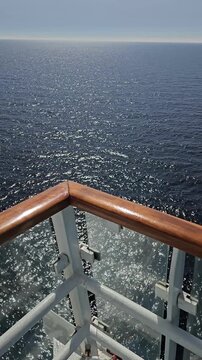Cruise ship deck overlooks vast blue ocean. Sunlight glints on choppy waters. White rail and polished wood handrail frame view. View from upper deck, calm seas. Horizon blends sky with distant ocean