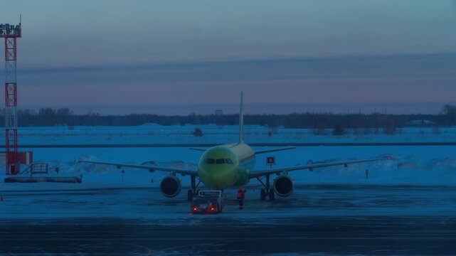 Passenger airplane towed by tractor on snowy airport apron at blue hour, winter dusk, warning lights, front view telephoto shot.