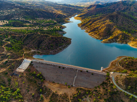 Aerial view of Dipotamos Reservoir with its dam structure and water tower surrounded by dry hilly terrain and sparse vegetation in Dimos Lefkaron, Larnaca, Cyprus.