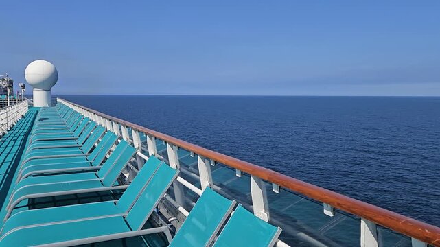 Cruise ship deck overlooks vast open ocean. Teal loungers line the railing for passengers to relax. Clear blue sky above calm sea horizon. White sphere fixture marks ship navigation point