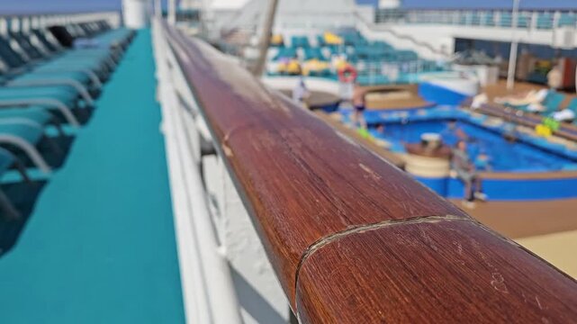 Wooden railing on cruise ship deck. Teal lounge chairs line the deck. Pool area visible in background. Clear blue sky overhead. Bright sunny day outside. Shallow focus on rail detail
