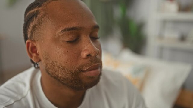 Man sitting on bed wearing a white tshirt, face in profile and eyes closed inside a building; quiet contemplation.