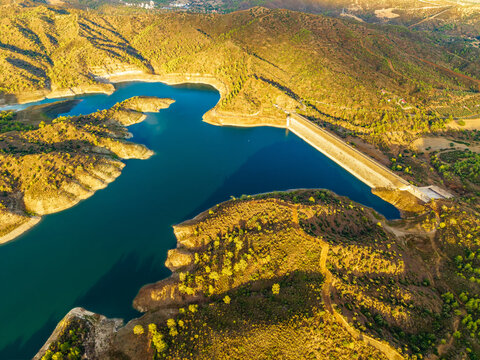 Aerial view of Dipotamos Reservoir surrounded by rugged hills, pine trees, and golden sunlight in Dimos Lefkaron, Larnaca, Cyprus.