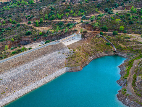 Aerial view of Dipotamos Reservoir featuring its turquoise water, stone dam wall, and surrounding hills with sparse vegetation in Dimos Lefkaron, Larnaca, Cyprus.