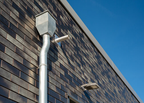 Metal downspout pipe mounted on dark ceramic brick facade under clear blue sky. Exterior drainage system detail suitable for architecture, construction and building design. Copy space